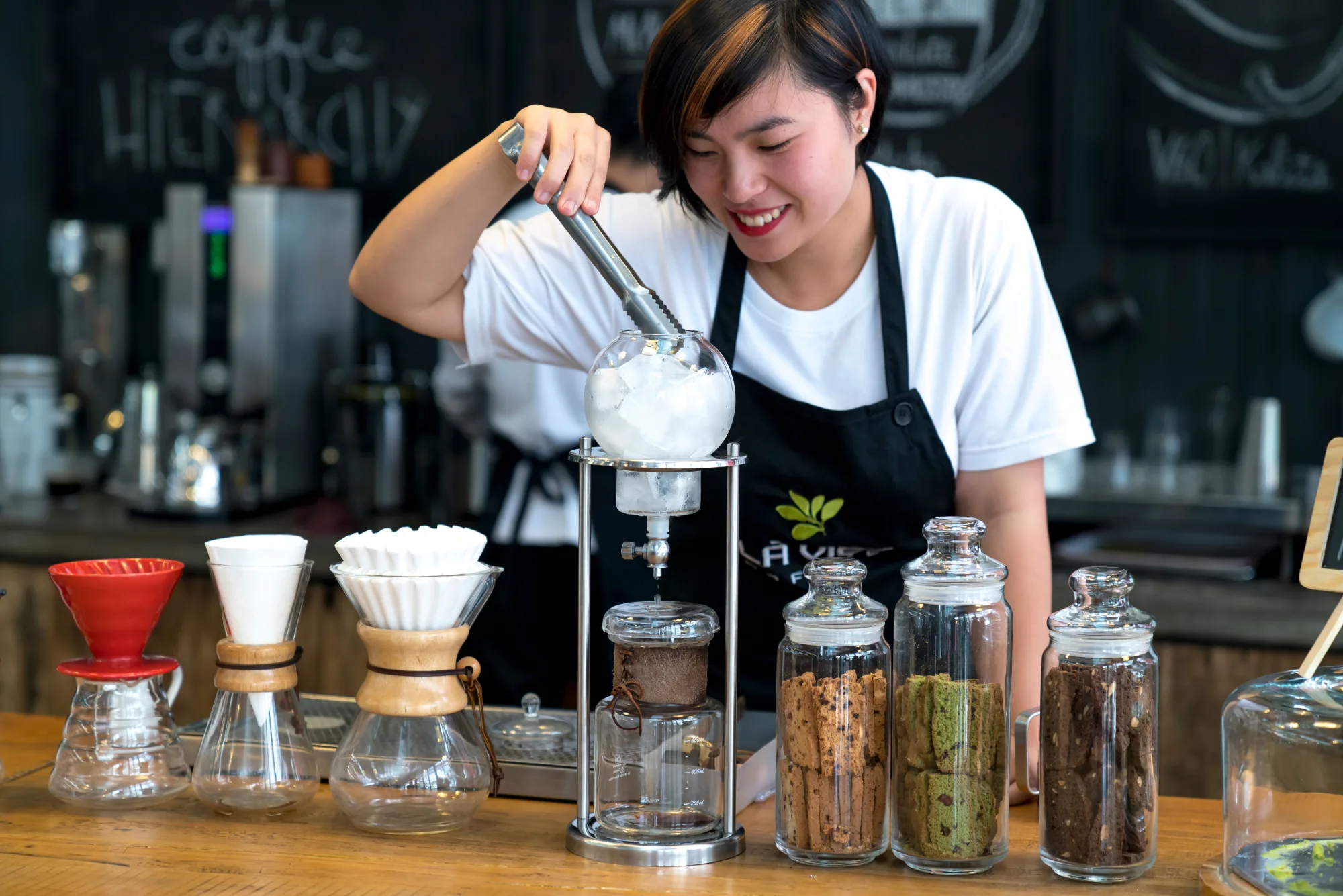 A barista actively working a siphon coffee brewer, filling the top chamber with ice