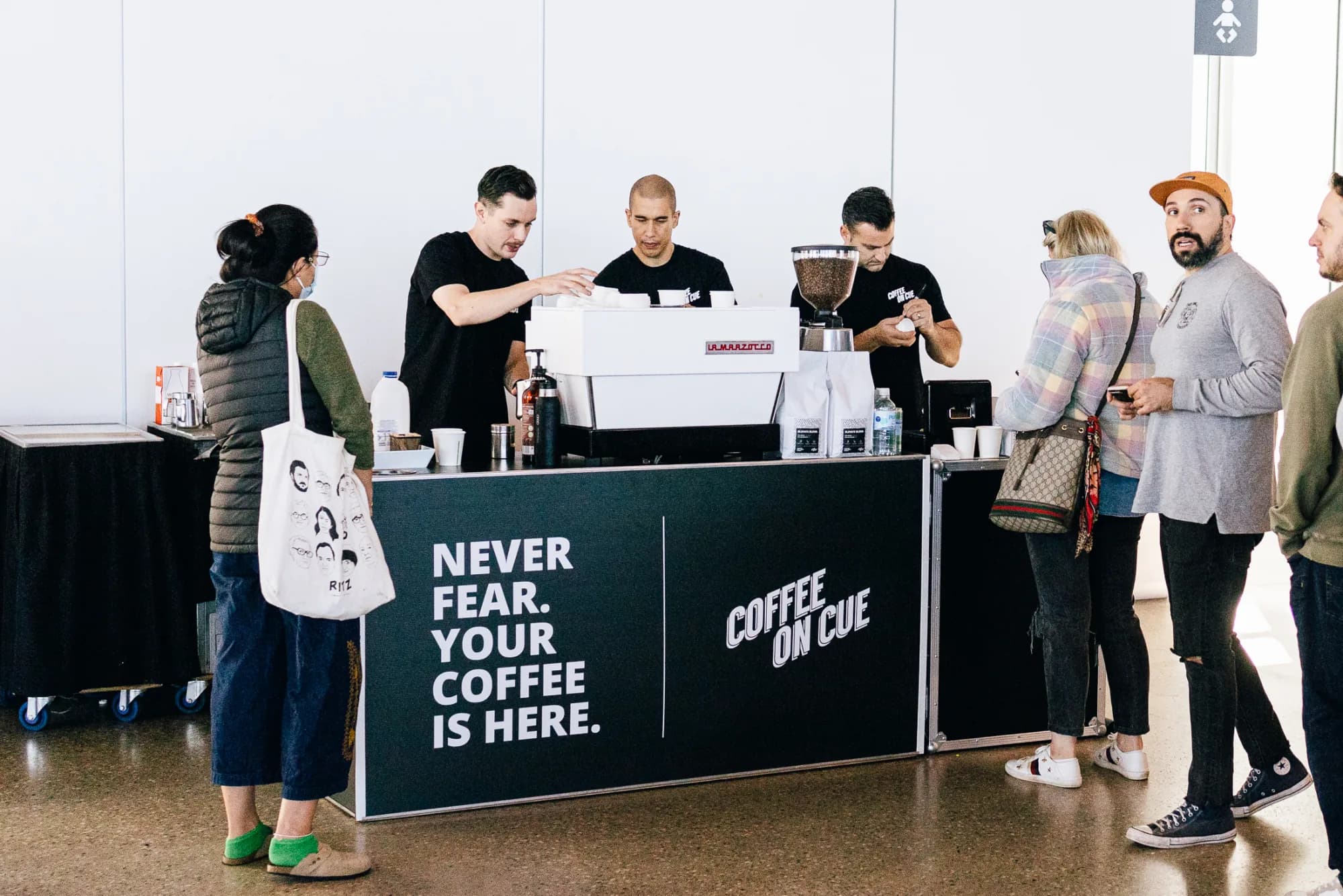 Three Coffee on Cue baristas working one branded cart with a La Marzocco machine while event guests wait on both sides of the counter