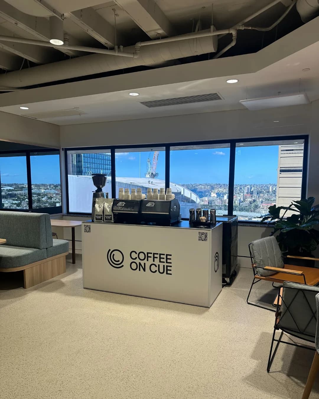 A Coffee on Cue cart set up in an office breakout space beside floor-to-ceiling windows with a city view, takeaway cups stacked on the machines