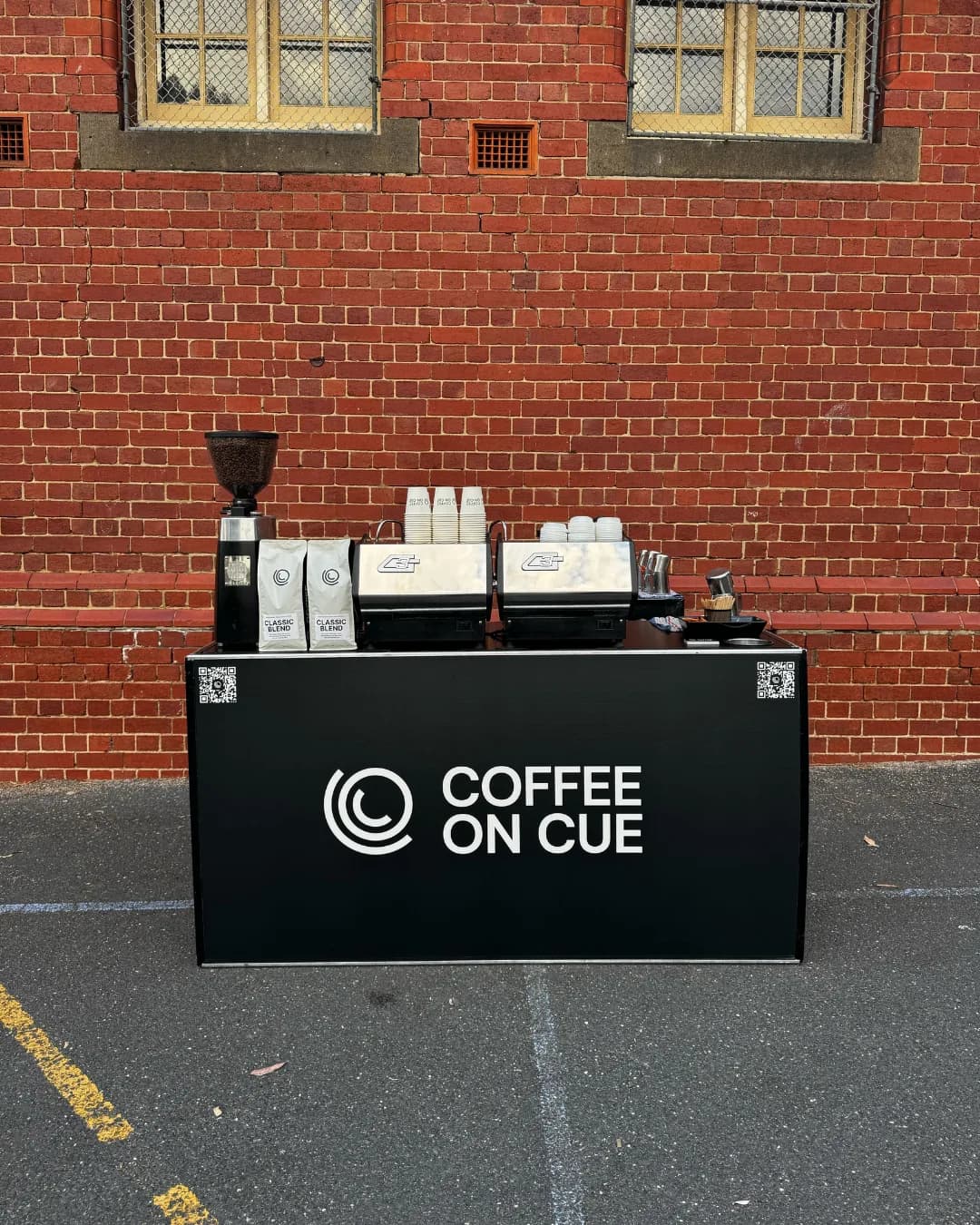 A Coffee on Cue cart set up against a red brick wall with a grinder, beans and espresso machine