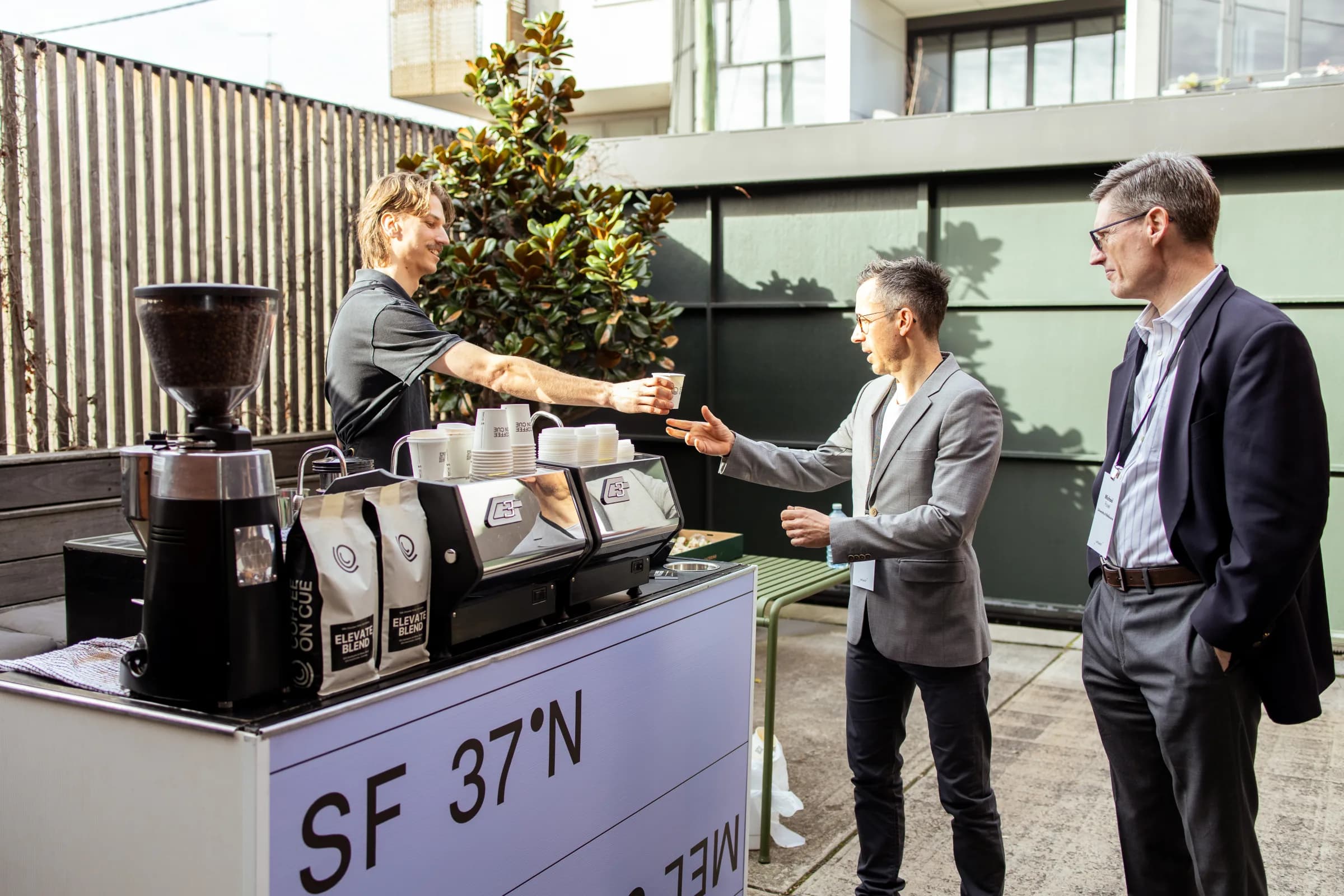 A Coffee on Cue barista handing a coffee across a client-branded cart to a guest at an outdoor corporate event
