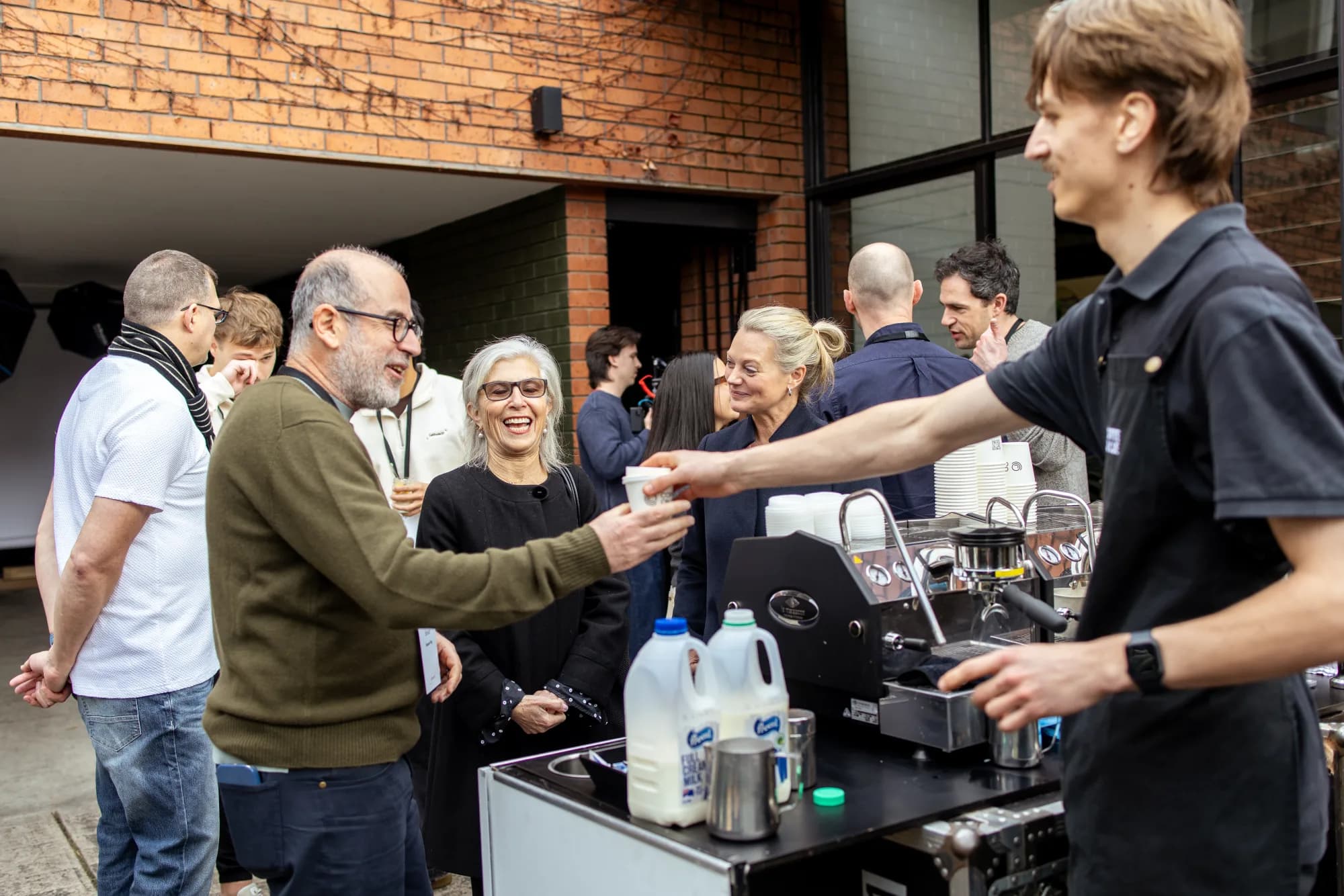 A barista handing a coffee to a guest in an outdoor Melbourne courtyard, a mixed-age group of guests around the cart