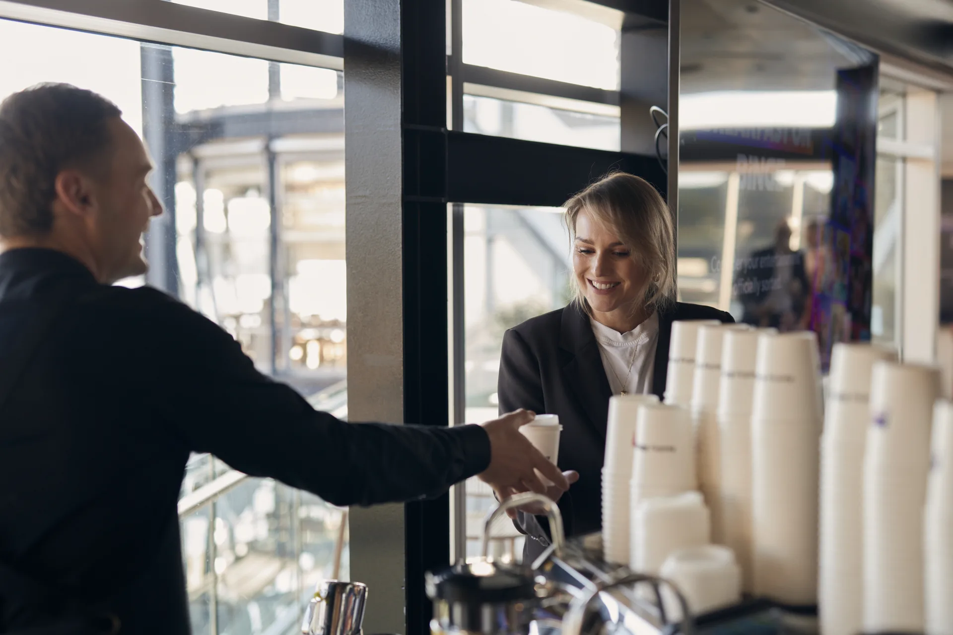 A Coffee on Cue barista in a black uniform handing a takeaway coffee across the counter to a smiling guest at a harbourside Sydney event, with a stack of cups in the foreground