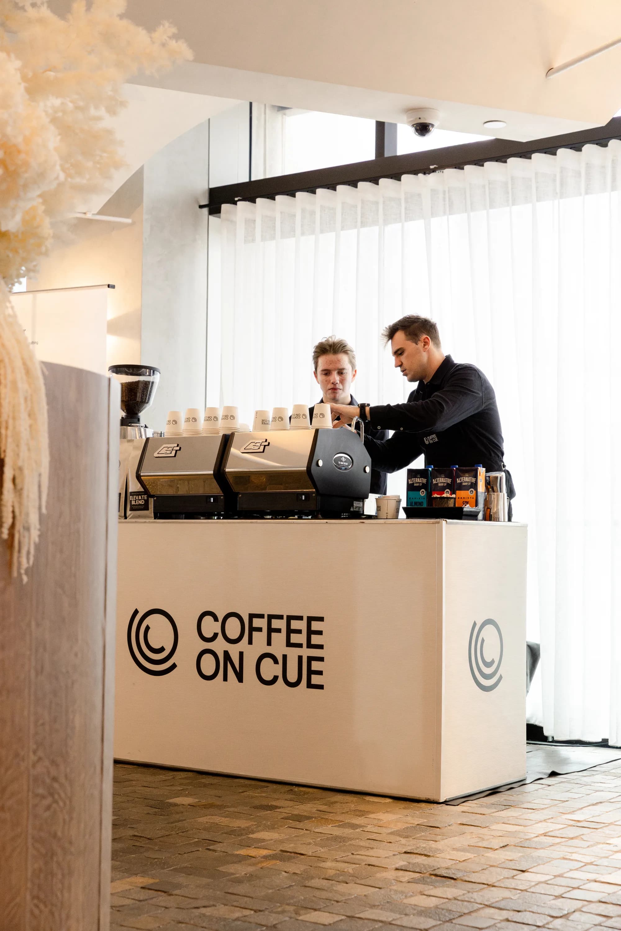 Two baristas preparing a La Marzocco machine at a white Coffee on Cue branded mobile coffee bar, with stacked cups and alternative milk cartons on the counter