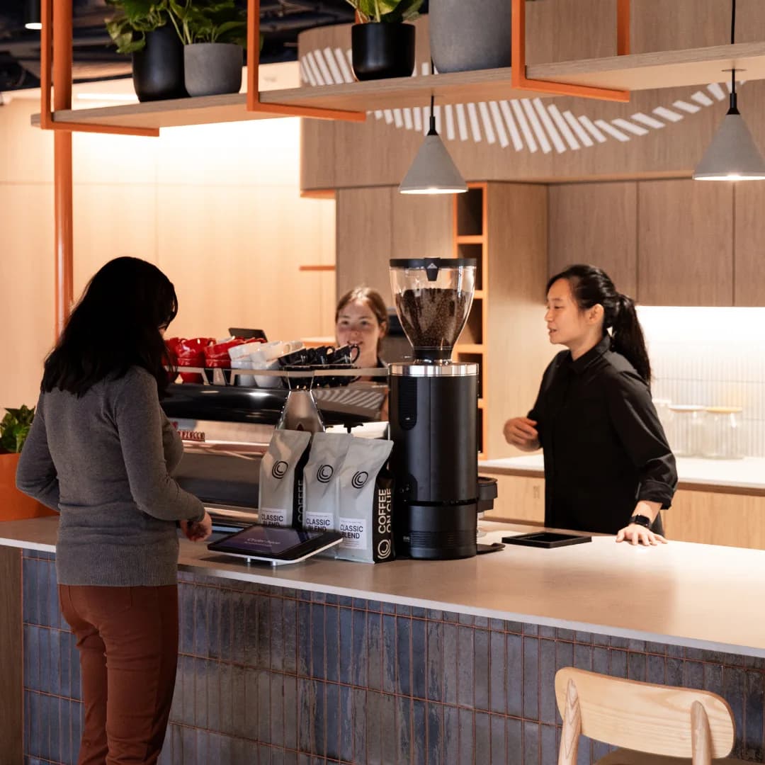 A colleague standing at a tiled workplace coffee counter while two baristas talk with her behind a grinder, bags of Classic Blend and an ordering tablet