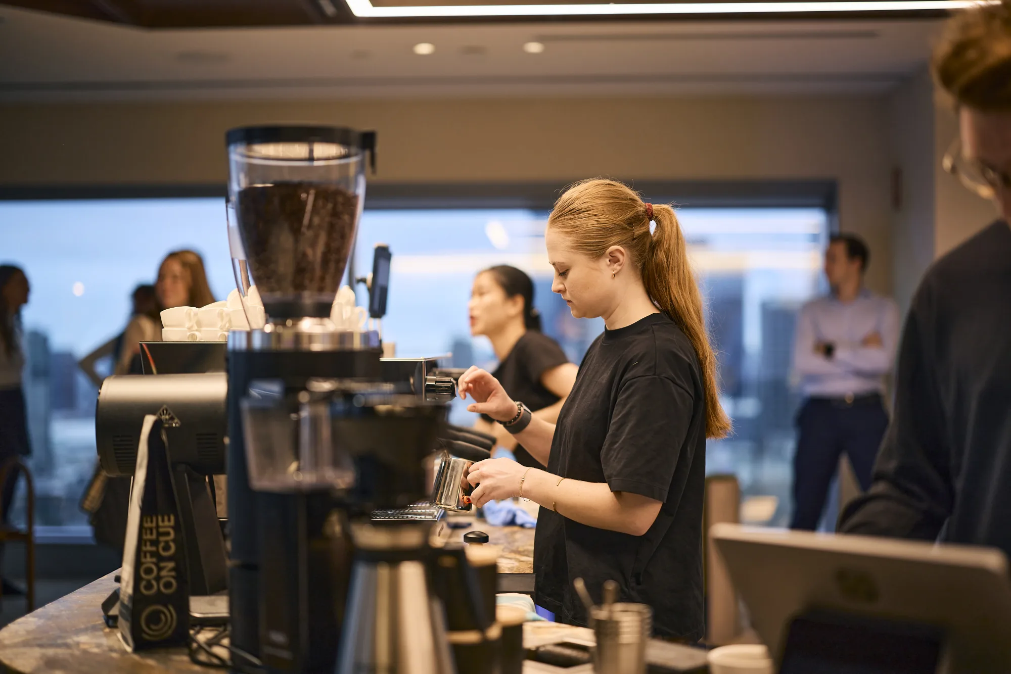 A Coffee on Cue barista working the grinder at the JLL 101 Collins bar, colleagues waiting beyond