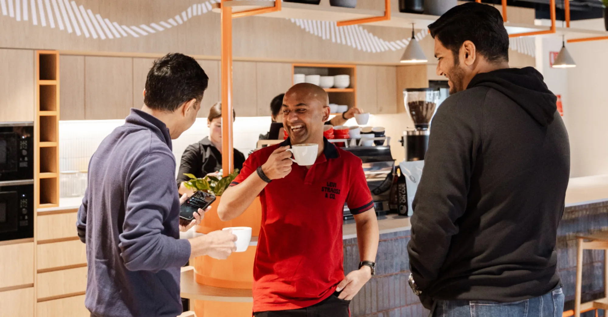 Three colleagues laughing together over coffee at a Coffee on Cue in-house office bar, a barista pouring behind them
